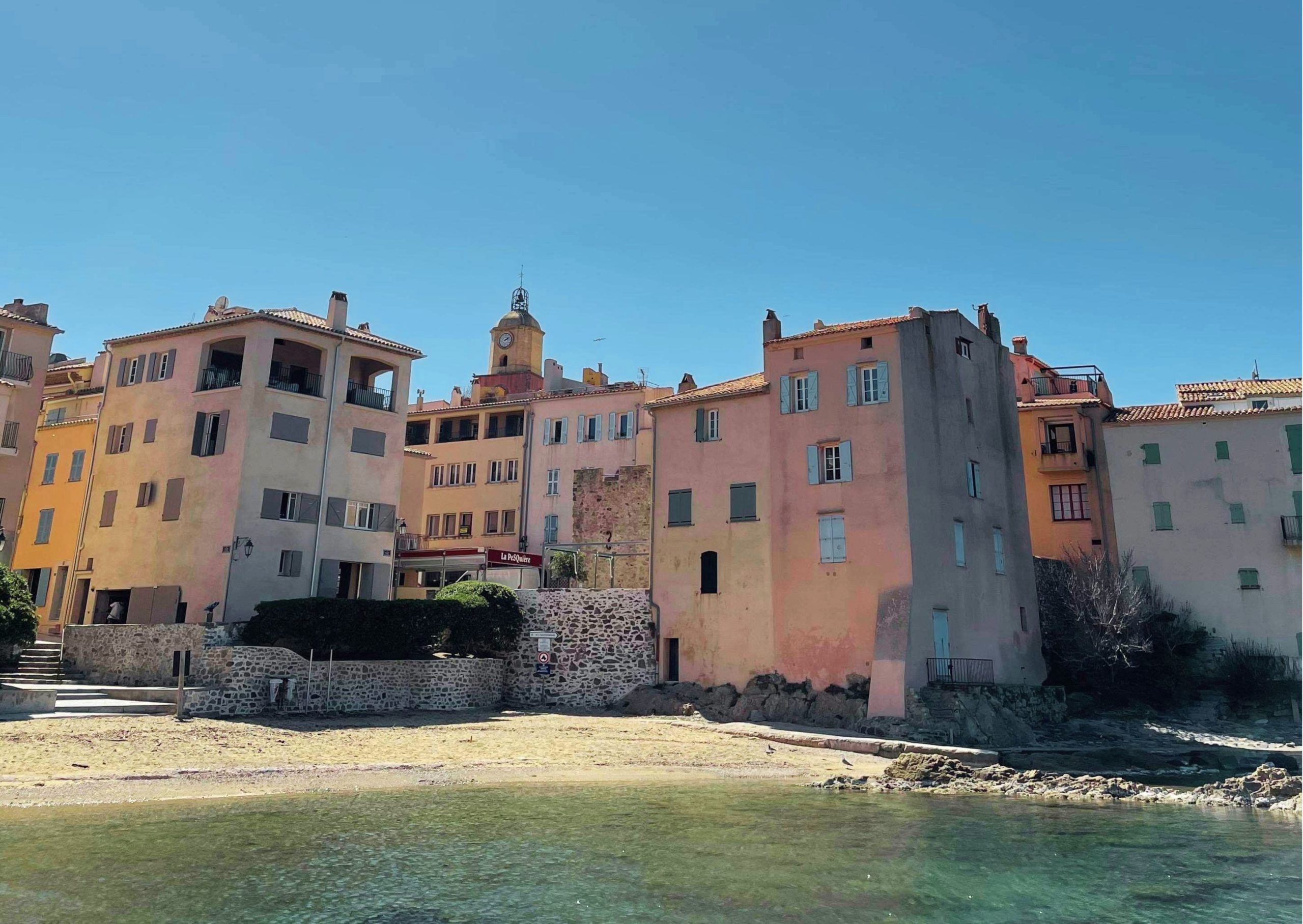Le clocher emblématique de Saint-Tropez se détache sur un ciel bleu, entouré des mâts de bateaux du port et des appartements colorés du village